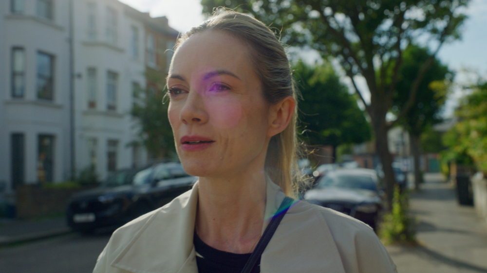 A close up shot of our campaign character Joanna. Joanna is wearing a cream coat, stood on the pavement in a suburban street with the sun shining down on her while looking out across the street.