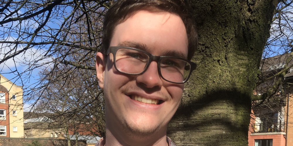 David Hogg is smiling, wearing a pink floral shirt and black glasses. He's stood in front of a tree trunk with grass, blue sky and buildings behind him.