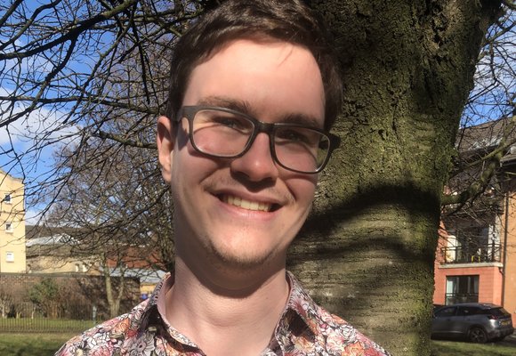 David Hogg is smiling, wearing a pink floral shirt and black glasses. He's stood in front of a tree trunk with grass, blue sky and buildings behind him.