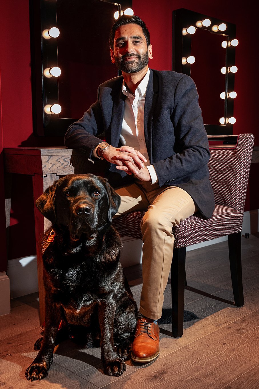 This portrait is of Amit, a man in his mid-forties with brown skin, short dark hair and a neatly trimmed dark beard, sitting in a warmly lit dressing room, gently smiling at the camera, accompanied by his black Labrador guide dog, Quark, seated proudly at his feet. Amit wears a smart navy blazer, white shirt, tan chinos, and polished brown leather shoes. His posture is relaxed yet composed, with his hands gently clasped and a confident, approachable expression on his face. The setting – with dark red walls, lit vanity mirrors behind him, and soft wood flooring – suggests a backstage environment. The lighting is soft and directional, casting warm highlights that flatter both Amit and Quark, creating a sense of focus. Quark, sitting tall and steady, looks directly at the camera, exuding calm and attentiveness. The pairing between the two speaks to trust, partnership, and shared independence. Altogether, it’s a powerful yet understated portrait of confidence, companionship, and quiet charisma.