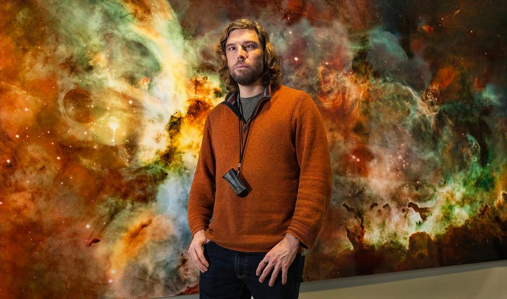 This portrait is of Nicolas, a man in his early forties with fair skin, standing confidently in front of a vast, glowing image of the Carina Nebula – a celestial cloudscape alive with swirling streaks of orange, green, blue, and red. As the nebula pulses with energy behind him, Nicolas wears a burnt-orange half-zip jumper over a dark top, paired with dark jeans. Around his neck hangs a black monocular – a tool Nicolas has been using since he was seven as an aid to help with distance vision such as crossing roads. His shoulder-length brown wavy hair and beard frame a calm, thoughtful expression in a scene that feels suspended where man and universe share the same space.