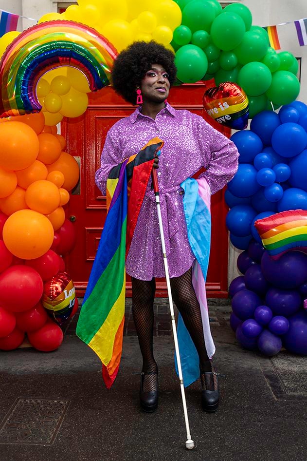 This portrait is of Ebony, a person with black skin sporting a bold black afro wig, proudly standing in front of a colourful balloon archway decorated for the London Pride celebrations. The background features a vivid red door surrounded by rainbow-coloured balloons, including a large foil rainbow balloon and heart-shaped balloons that say, “Love is Love.”

Ebony is dressed in a sparkly, purple sequin shirtdress, fishnet tights, and black heels. They are wearing bright pink rectangular earrings and have striking makeup on with pink eyeshadow. They confidently pose with one hand on their hip and the other holding a white cane. Multiple Pride flags – including the traditional rainbow flag, the Progress Pride flag, and the transgender flag – are draped around them, symbolising inclusivity and intersectionality.

This vibrant image is both joyful and empowering, celebrating LGBTQ+ pride, visibility, and diversity with a strong sense of self-expression and inclusion.