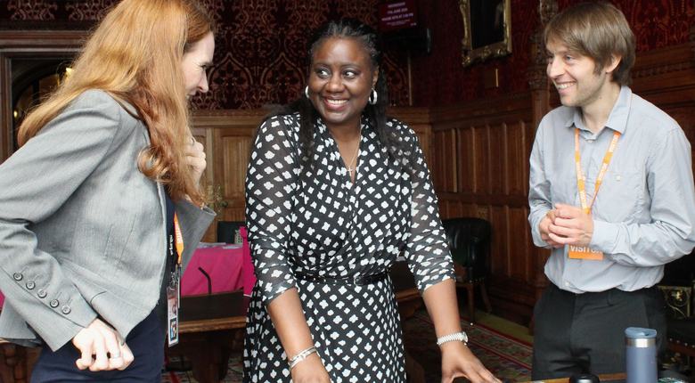 Marsha de Cordova MP stands behind a table flanked by two RNIB staff members. They are all smiling and in conversation. The room is very fancy and ornate, richly decorated with wood panelling and red patterned wallpaper. The table they stand around features a McGonagle Reader, which is an audio and tactile accessible voting device.