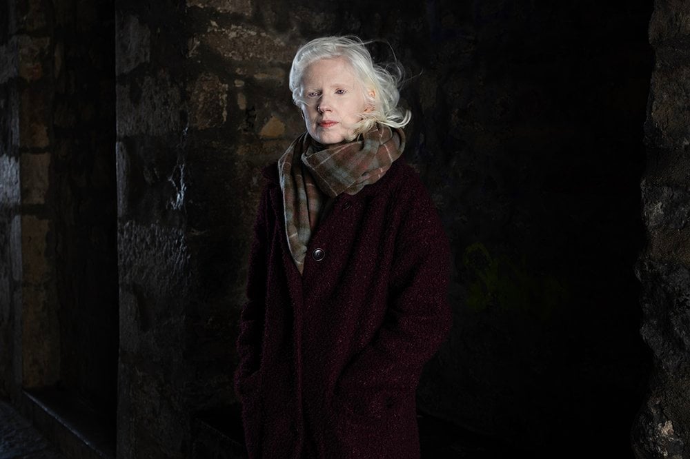 This portrait is of Ellen, a woman with fair skin and fair hair, standing against a dark, old stone-walled background. The lighting is soft and moody, casting gentle shadows that give the image a quiet, almost timeless quality. Ellen’s fair hair and skin catch the available light, making her look luminous. She is wearing a deep burgundy textured coat and a large, cozy tartan scarf in muted earthy tones, wrapped warmly around her neck. Her hands are tucked into her coat pockets, and she looks directly at the camera with a calm, steady gaze. The overall mood of the striking portrait is contemplative and elegant, blending natural light with rich textures and heritage tones, evoking a deep sense of place, history, and quiet strength.