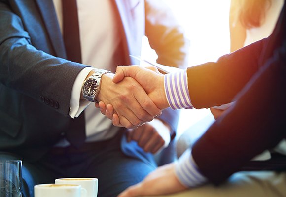 Two people dressed in suits, shaking hands across a table.