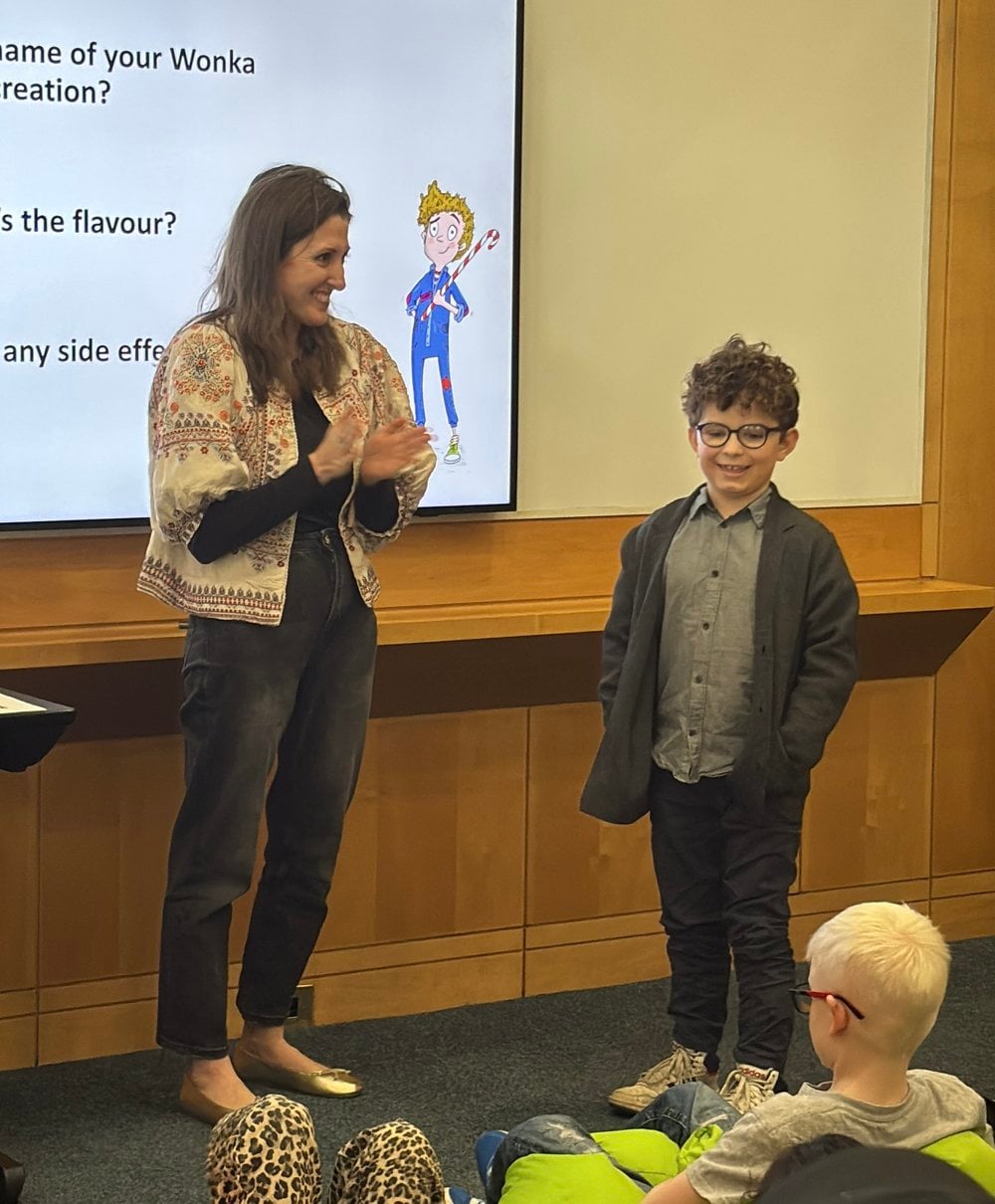 Ernest Mozakka is standing at the front of the class smiling with Sibéal Pounder (whose book was part of the Work Book Day 2026 lineup). Sibéal is clapping beside him and announcing Ernest as the winner of RNIB's 2026 creative writing competition.