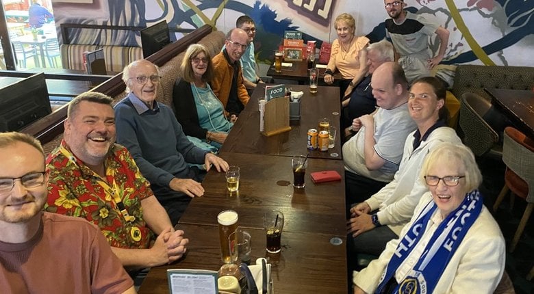 Football Focus, an RNIB Scotland weekly telephone group, all meet together. A group of 12 people sat around a table in a restaurant smiling. Jean is bottom right, smiling and wearing a football scarf.