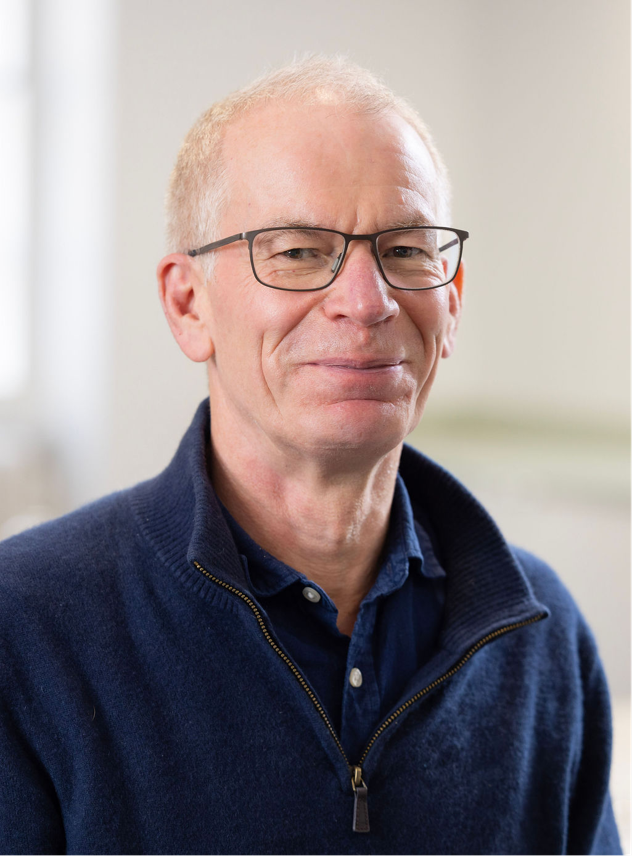 A picture of Graham Colbert, a man with short grey hair and glasses wearing a dark blue shirt and dark blue quarter zip jumper.