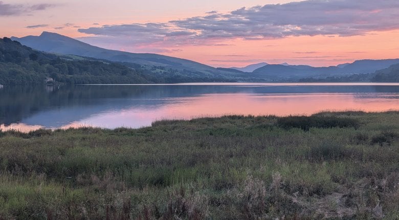An image of the Welsh country side. The view is of a lake at sunset, the sky is a dusty orange and blue colour.