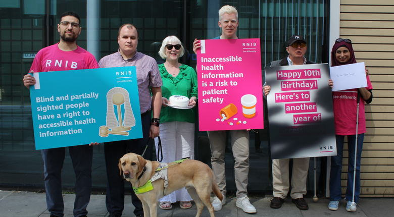 A group of six RNIB supporters stand outside the Department of Health and Social Care with placards to mark the 10th Anniversary of the Accessible Information Standard. One sign reads, “Blind and partially sighted people have a right to accessible health information,” while another states, “Inaccessible health information is a risk to patient safety.” A guide dog stands in the foreground. Another placard celebrates “Happy 10th birthday, here's to another year of" with the remaining words blacked out to show the impact of diabetic retinopathy on reading information.