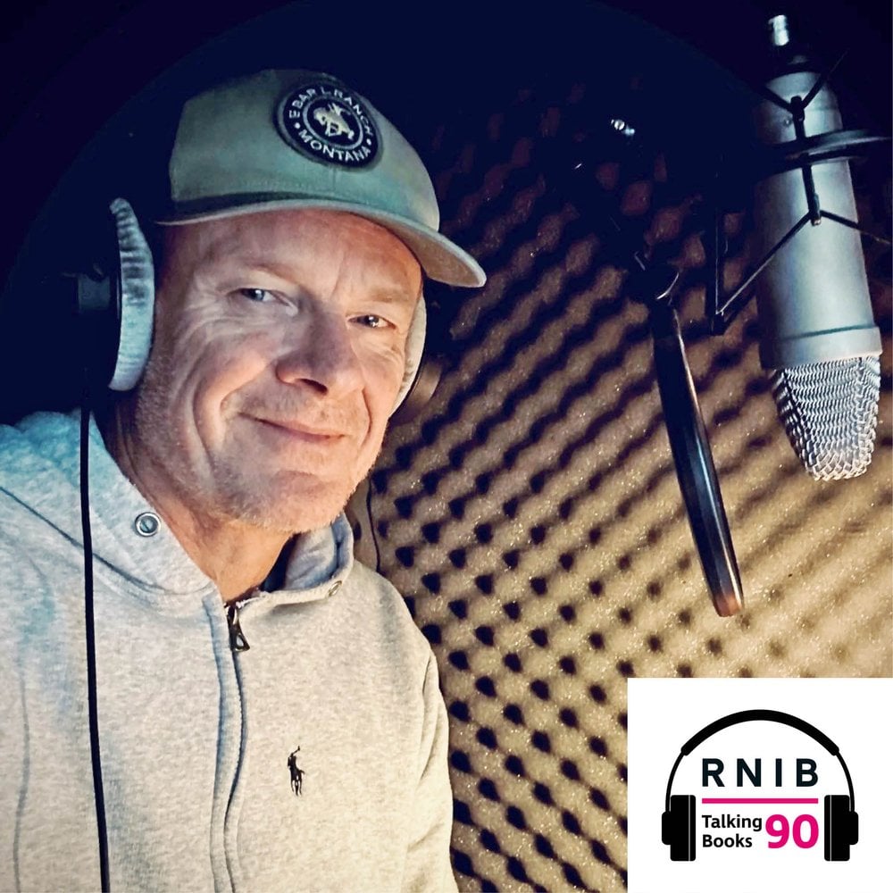 Alistair Petrie, a man in his mid 50s standing in a recording booth in front of a microphone, smiling and looking at the camera. He is wearing a grey hoodie, a grey baseball cap and over ear headphones.