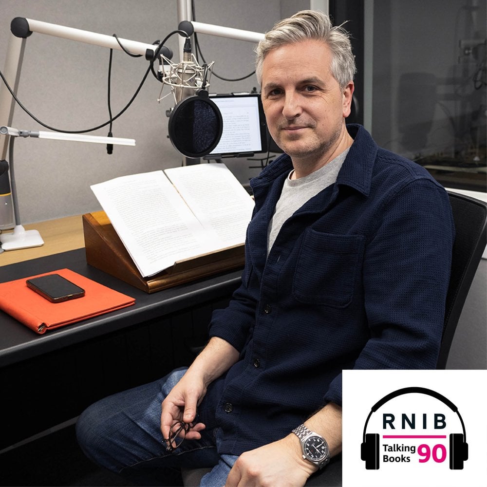 Ben Willbond, a man in his early 50s sat in a recording studio, turned to face the camera. He is wearing a navy blue cardigan and blue jeans.