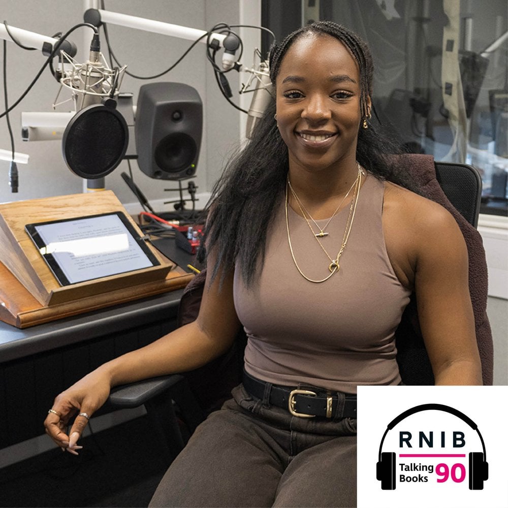 Corrina Brown, a woman in her late 20s sitting on a chair in a recording studio smiling facing the camera, she is wearing a light brown tank top and dark brown jeans.