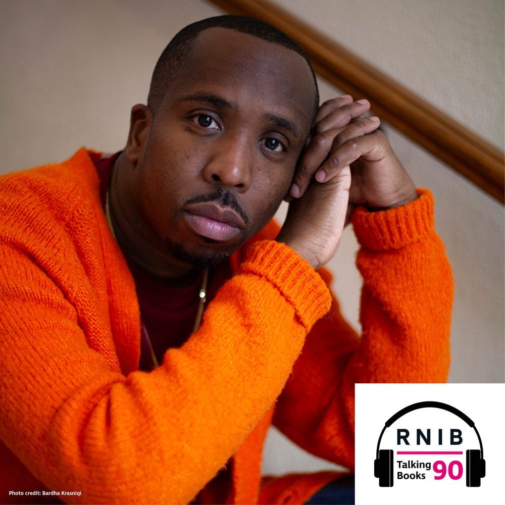 Kiell Smith-Bynoe, a man in his mid 30s sitting on the stairs looking at the camera, he is wearing an orange cardigan.