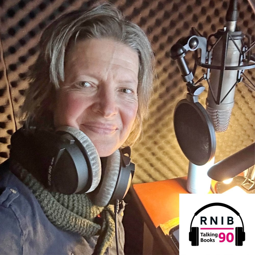 Lucy Scott, a woman in her mid 50s in a recording booth turned to face the camera smiling, a microphone is in front of her. She is wearing a dark blue shirt, olive green scarf and over ear headphones around her neck.
