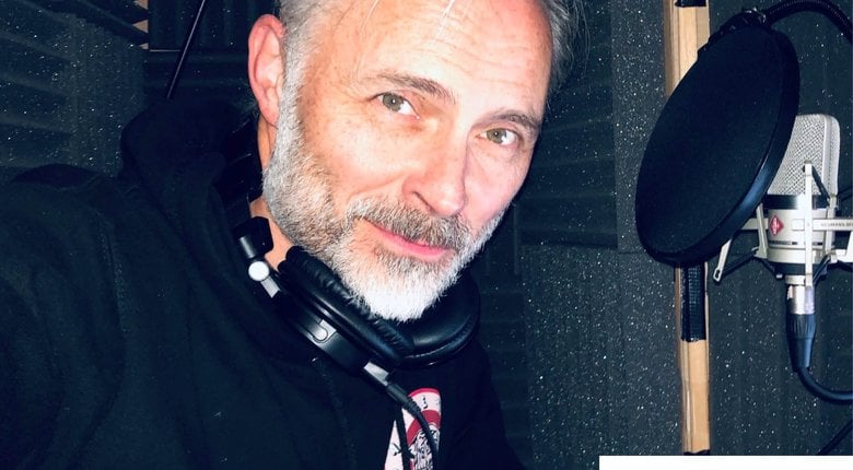 Mark Bonner, a man in his late 50s standing inside a recording studio in front of a microphone, he is turned to face the camera smiling. He is wearing a black hoodie and over ear headphones around his neck.
