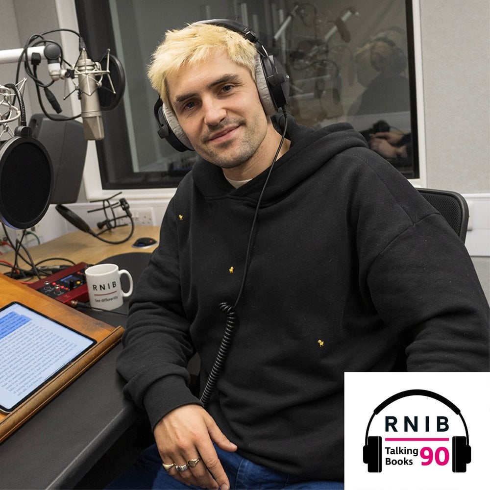 Phil Dunster, a man in his early 30s sitting in a chair in a recording studio whilst smiling and looking at the camera. He is wearing a black hoodie, blue jeans and large over ear headphones.