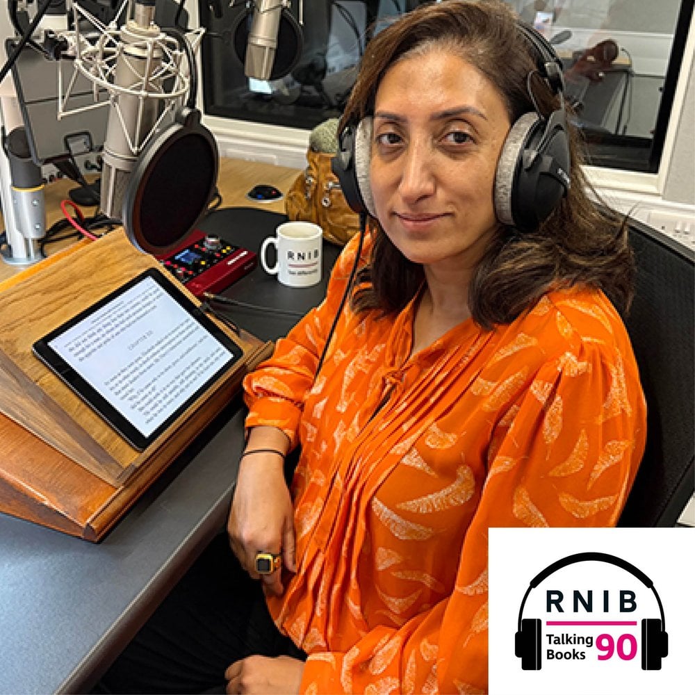 Shazia Mirza, a woman in her early 50s sitting in a recording studio, smiling at the camera whilst wearing an orange top and headphones.
