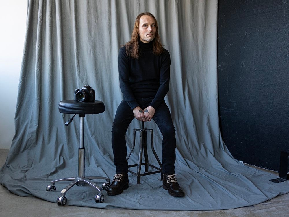 Ian, a man in his mid to late forties with fair skin, sitting calmly on a metal high stool in a studio setting. With long, light brown, slightly wavy hair that falls past his shoulders, Ian is dressed entirely in black with a fitted turtleneck jumper, slim jeans, and sturdy lace-up boots. His hands are loosely clasped between his knees, revealing a sparrow tattoo on the back of each hand. . Additionally, he has a small geometric tattoo on his right index finger that looks like a series of downward-pointing arrows stacked vertically – these are dream catcher symbols. Ian’s head is slightly to the side while his gaze is to the camera with a thoughtful, somewhat introspective expression.
Next to him is another stool with his camera resting on top — a nod to his work. The background is a soft grey drape that pools naturally onto the floor, creating a quiet, intimate atmosphere. The overall tone of the minimalist portrait is artistic and contemplative conveying creativity and solitude.