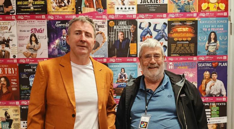 Jamie MacDonald in an orange suit and holding his white cane, with our volunteer Bill, both standing in front of a wall of Fringe posters
