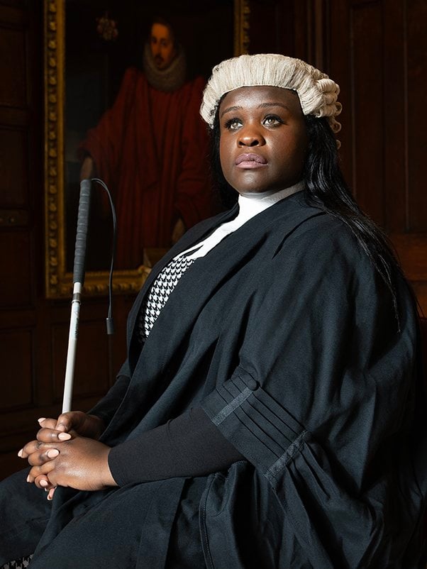 This portrait is of Jessikah, a woman in her mid-twenties with black skin, dressed in formal barrister attire – she wears a traditional white wig and a black robe over a white top and checkered vest. She holds a white cane and her expression is one of confidence and poise.
The background is richly decorated with dark wood panelling and a portrait of Henry Montagu, 1st Earl of Manchester, looking over her as she poses in the Queen’s Room at the Honourable Society of the Middle Temple in London. The overall composition suggests strength and professionalism.