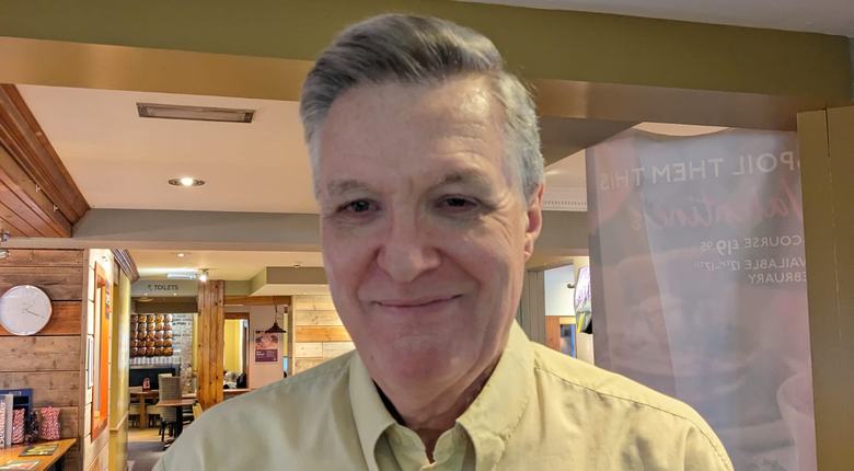 John, RNIB volunteer, stands smiling at the camera. He has short grey hair and and is wearing a light yellow shirt.