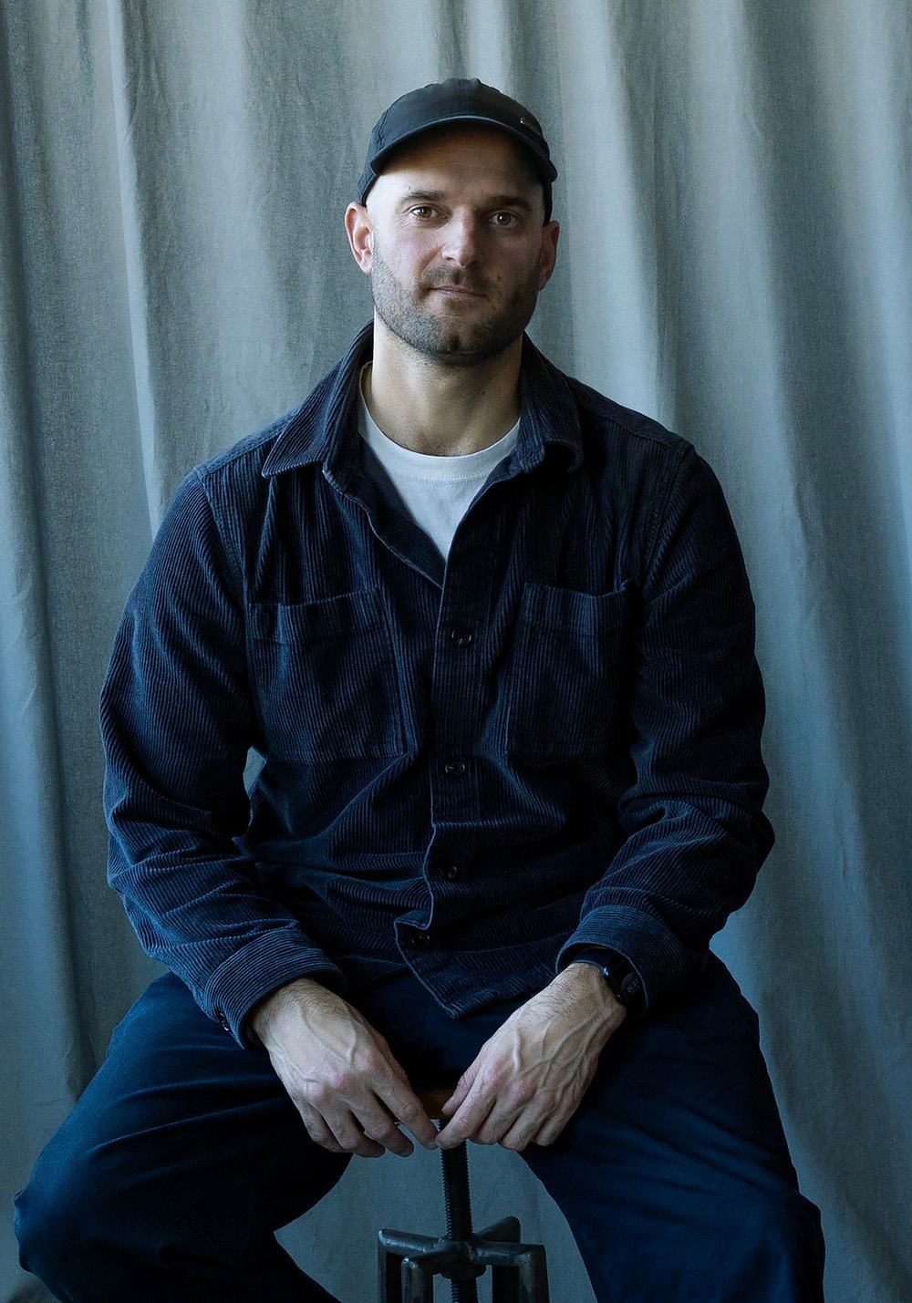A man wearing blue corduroy hat, shirt and trousers sitting on a stool against a white sheet background