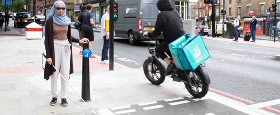 Khadija stands next to a shared space sign on the pavement of Pentonville Road. A e-bike zooms past her left hand side into the shared space