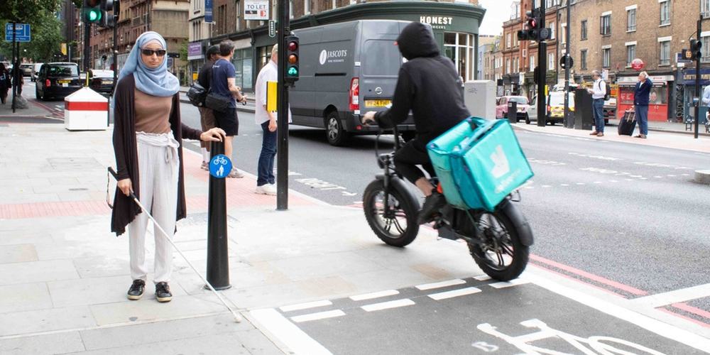 Khadija stands next to a shared space sign on the pavement of Pentonville Road. A e-bike zooms past her left hand side into the shared space