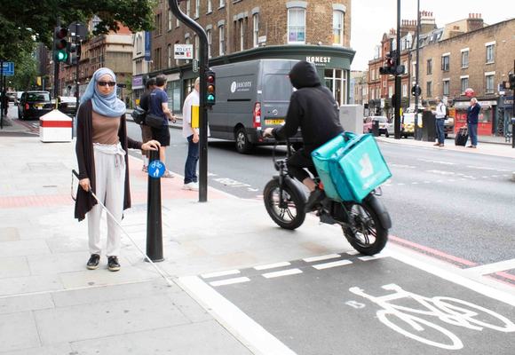Khadija stands next to a shared space sign on the pavement of Pentonville Road. A e-bike zooms past her left hand side into the shared space