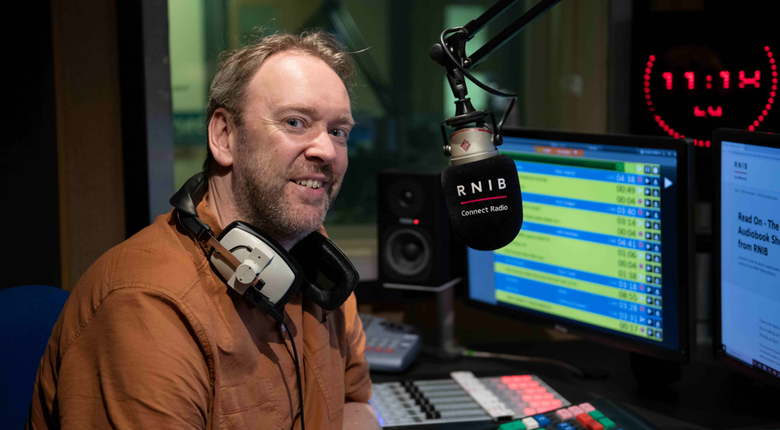 Kris sitting in the radio recording booth, smiling towards camera. In front of him is an RNIB Connect Radio microphone and sound desk.