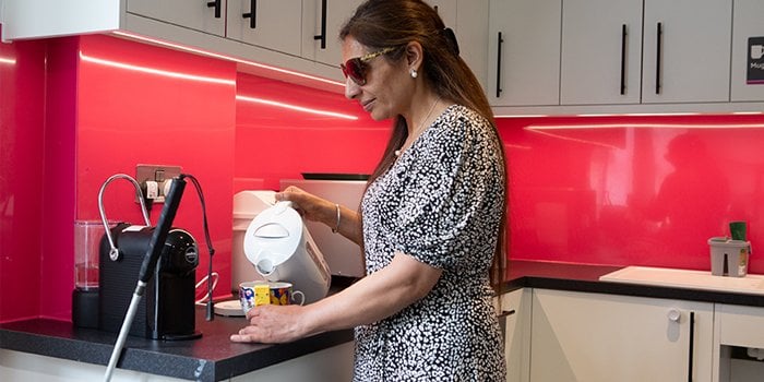 A woman with sight loss wearing a black dress with a white pattern, making a hot drink with a liquid level indicator, in an RNIB office kitchen.