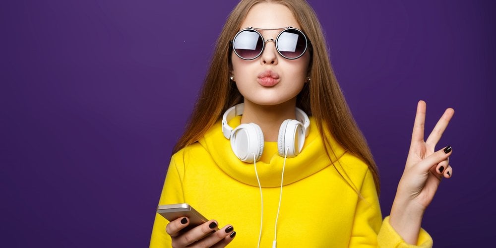 Lady in bright yellow dress wearing round sunglasses and white headphones round her neck connected to her phone in one hand. She is making a peace sign with the other hand.