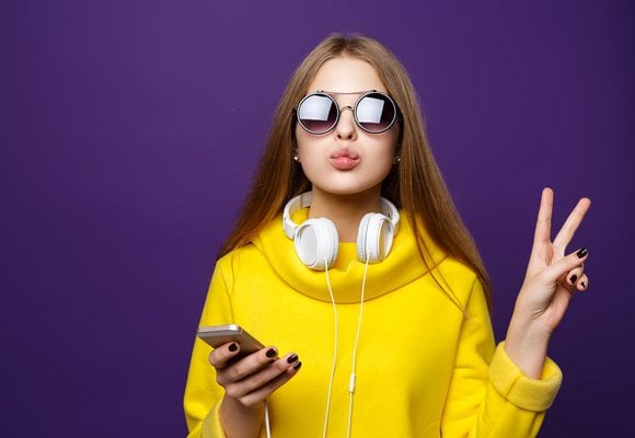 Lady in bright yellow dress wearing round sunglasses and white headphones round her neck connected to her phone in one hand. She is making a peace sign with the other hand.