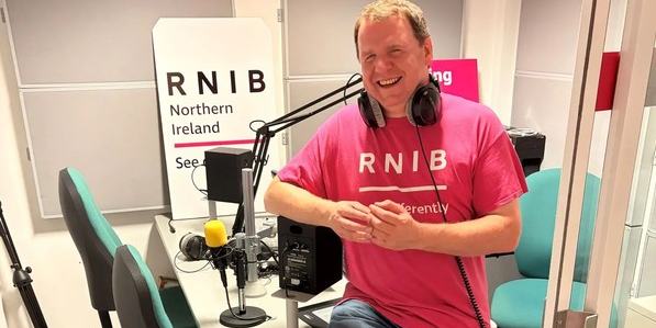 Joe Kenny sitiing on the edge pf the studio desk. He is wearing a pink RNIB T-shirt and is smiling at the camera.