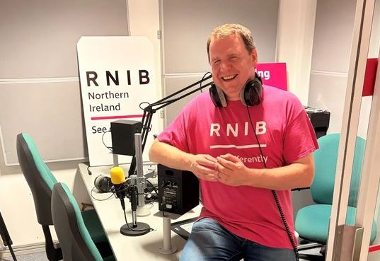 Joe Kenny sitiing on the edge pf the studio desk. He is wearing a pink RNIB T-shirt and is smiling at the camera.