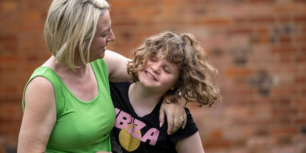 A mum with neck length blonde hair is wearing a green vest while hugging her young daughter, who is smiling and wearing a black top while flipping her brown wavy hair.