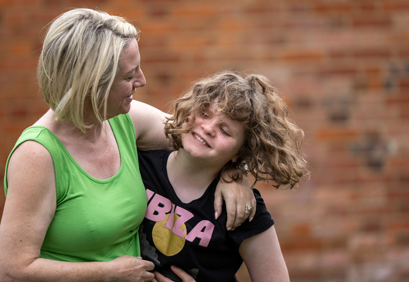 A mum with neck length blonde hair is wearing a green vest while hugging her young daughter, who is smiling and wearing a black top while flipping her brown wavy hair.