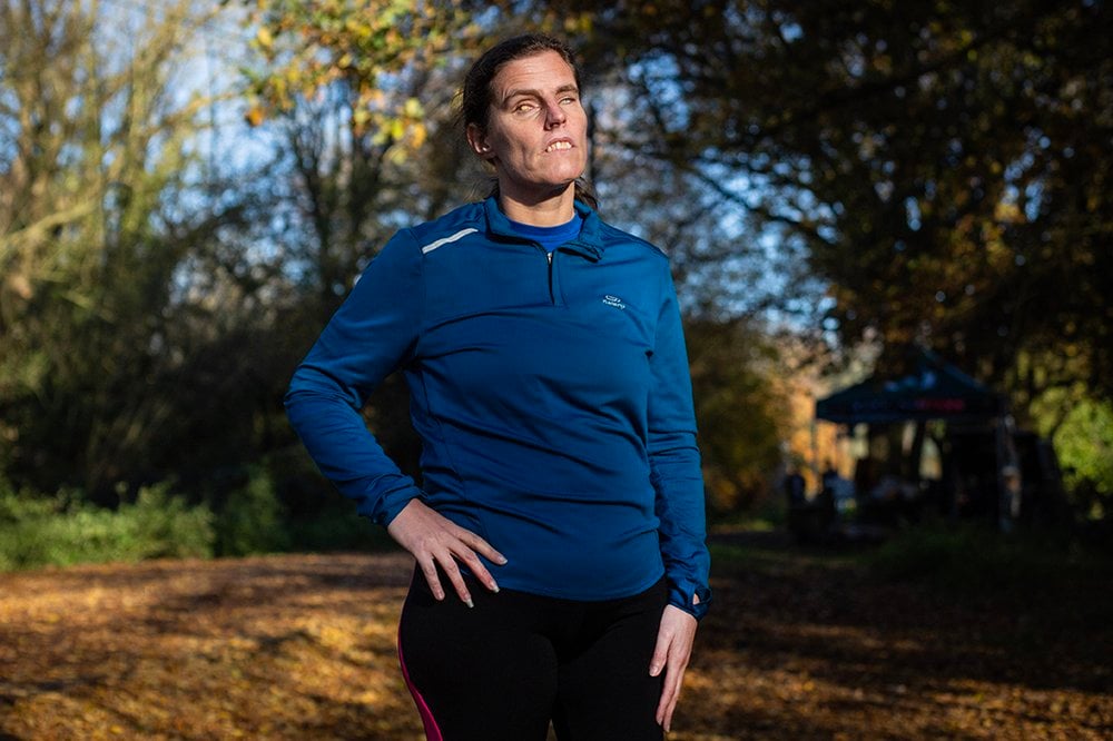 This portrait is of Louise, a woman in her late thirties with fair skin and dark pulled back hair, standing outdoors – the ground is covered with fallen autumnal leaves. She wears a blue long-sleeve athletic top and black leggings with a red stripe. She stands with her right hand on her hip, suggesting a moment of rest after running a marathon at Rickmansworth Aquadrome in Hertfordshire. Sunlight casts bold shadows that sculpt her face and upper body with warmth and clarity. Behind her, a wooded landscape unfolds – trees in varied hues, some tinged with golden leaves, signalling the shift of seasons. The setting suggests a calm and natural feel.