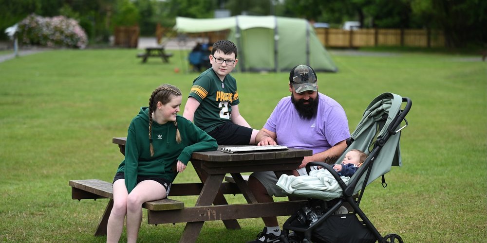 A man and two children (boy and girl) sit around a picnic table at a park, with a baby in a pram beside the man.