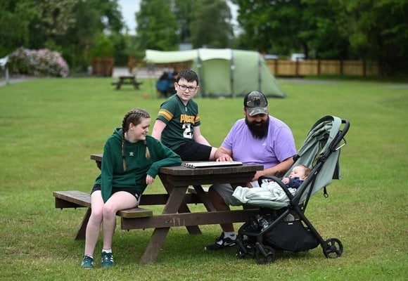 A man and two children (boy and girl) sit around a picnic table at a park, with a baby in a pram beside the man.