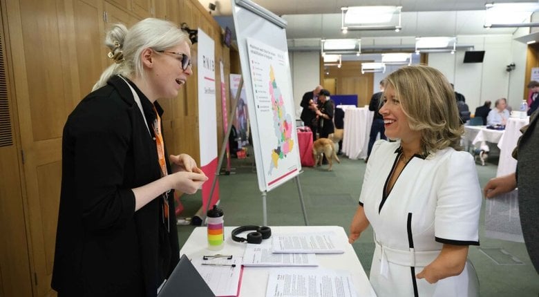 Alt text:   Marie Tidball MP and Hannah from RNIB stand talking at a table with documents and a laptop at an indoor event."