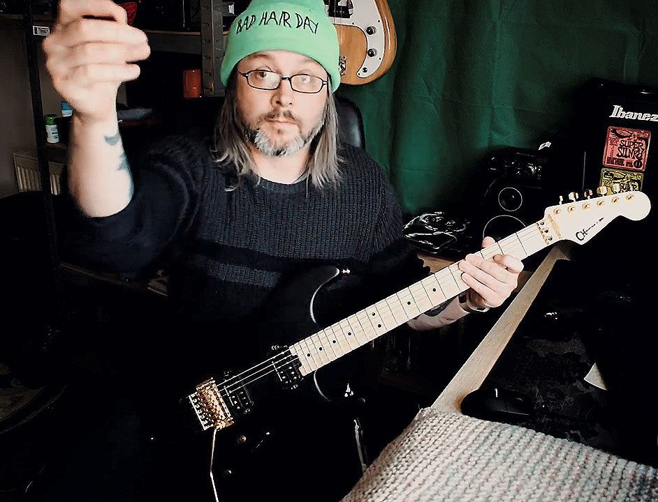 Mark Adams, RNIB volunteer, has shoulder length hair and a beard. Mark is holding a black electric guitar and is wearing a green beanie hat.