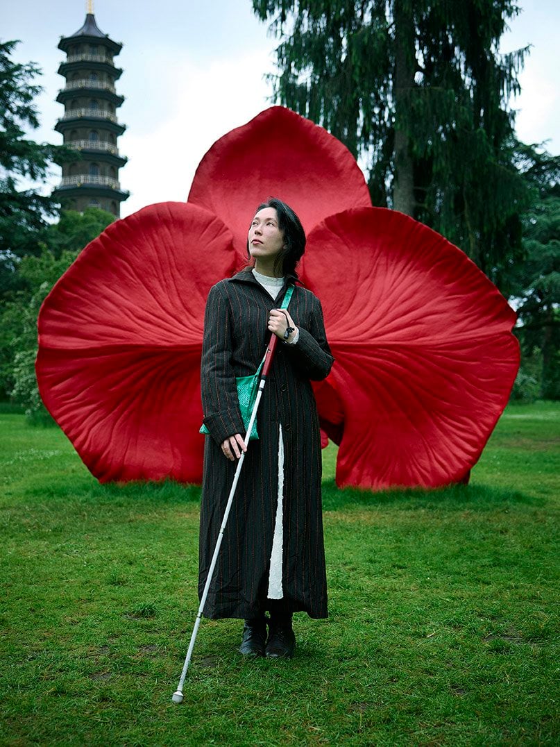 Maud, a woman in her late twenties with fair skin and long dark hair, posing outdoors at Kew Gardens in London, in front of a large, vivid red sculpture resembling flower petals by British visual artist Marc Quinn. She is wearing a long, dark pinstripe coat over a white garment and black boots, proudly holding her white cane. She also carries a green textured bag tucked under one arm.
Her gaze is directed slightly upward and to the side, suggesting an introspective, thoughtful expression. The bright red petals create a dramatic and bold backdrop, while a tall pagoda tower and lush green trees surround the scene behind her adding a serene and culturally rich atmosphere to the composition. The overall effect suggests quiet strength, elegance, and individuality.