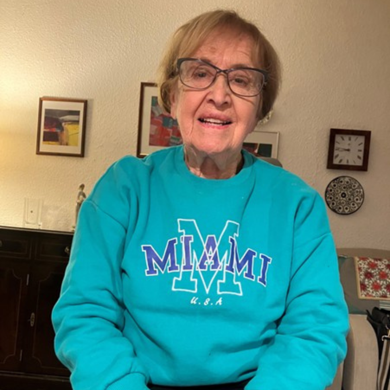 Image description: Mavis is smiling: she has short dark blonde hair and glasses. She is wearing a blue jumper and black trousers