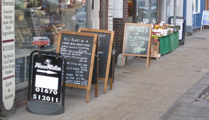 A row of shops along a street, all with pavement signs and advertising boards in front of them.