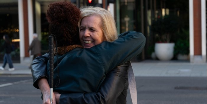 A white woman with blond hair on a city street holding a cane smiling and hugging another woman with brown hair and a black leather jacket who is facing away from the camera.