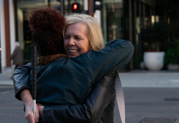 A white woman with blond hair on a city street holding a cane smiling and hugging another woman with brown hair and a black leather jacket who is facing away from the camera.