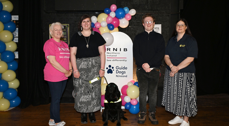 Four people stood with a black labrador dog against a black wall with multi-coloured balloons.