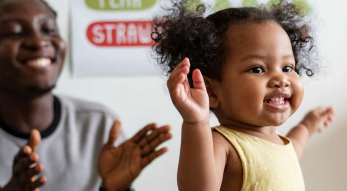 A parent claps and smiles as a toddler walks.
